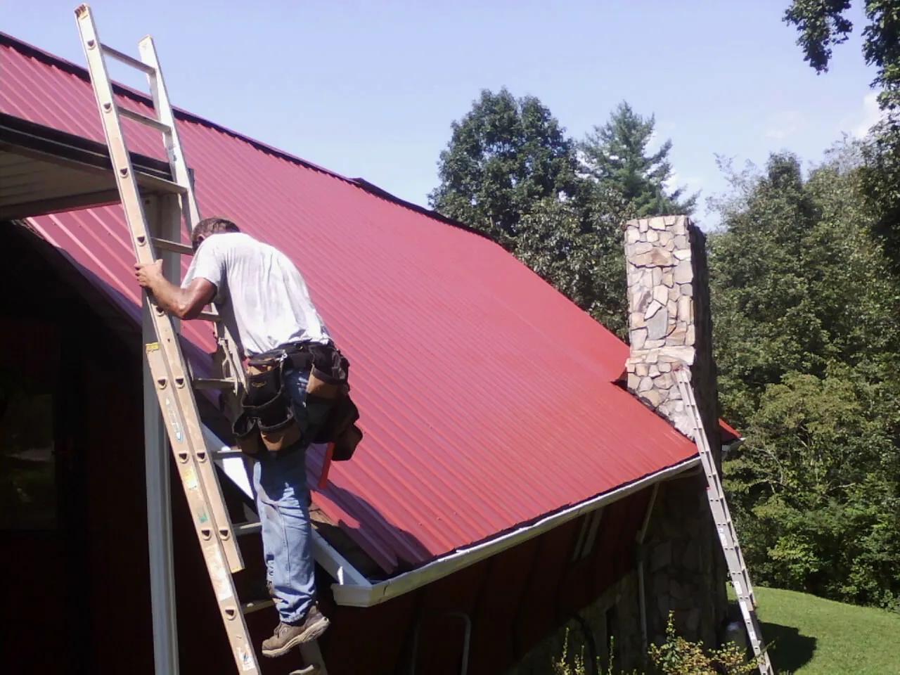 Roofer on a ladder beside a red metal roof and stone chimney — Soffit & Fascia Repair by Roofing Brothers in Churchton